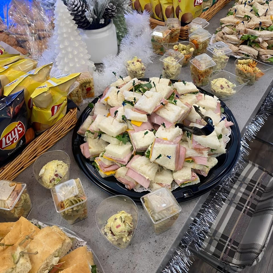 A festive buffet table with various sandwiches, pasta salad cups, and potato salad cups.
