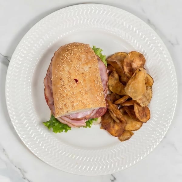 A deli sandwich with ham, salami, and lettuce on a sesame seed bun, served with potato chips.