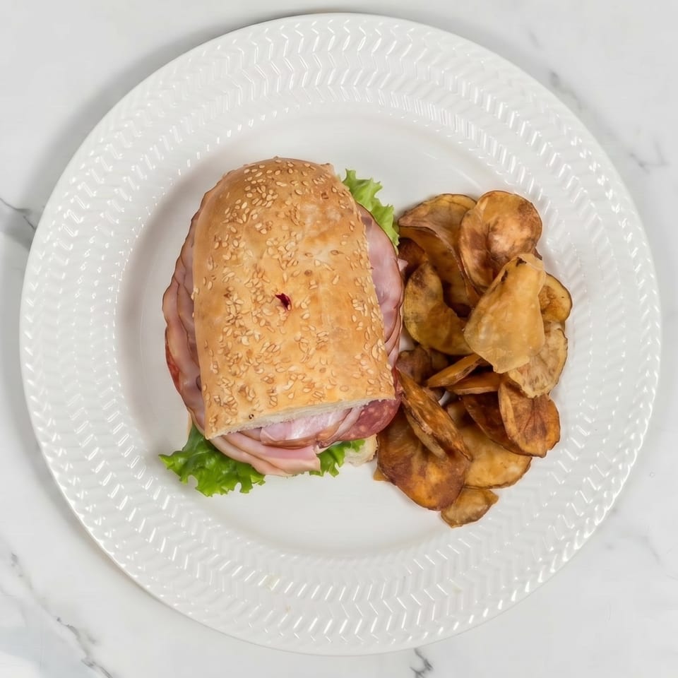 A deli sandwich with ham, salami, and lettuce on a sesame seed bun, served with potato chips.