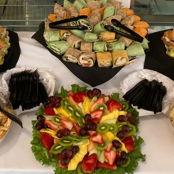 A catering spread with colorful wraps, a fresh fruit platter, and plastic cutlery.