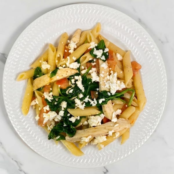 Penne pasta with chicken, spinach, diced tomatoes, and crumbled feta cheese on a white plate.
