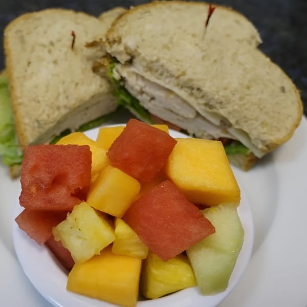 Turkey sandwich on wheat bread with lettuce and tomato, served with a side of mixed fruit.